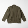 An olive green waffle-knit open-front cardigan sweater hanging on a black hanger against a textured off-white wall.