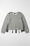 A heather gray long-sleeve top with dark gray shoulder panels and a chest pocket, hanging on a black hanger against a textured off-white wall. A white shirt is layered underneath at the hem.