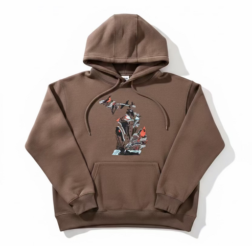 A brown pullover hoodie with a graphic print of birds and branches, laid flat on a beige branded paper surface over a wooden floor.