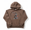 A brown pullover hoodie with a graphic print of birds and branches, laid flat on a beige branded paper surface over a wooden floor.