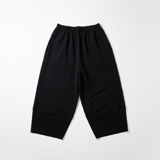 Black wide-leg cropped pants hanging on a black hanger from a wall rack, with a light-colored textured wall and wooden bench visible in the background.