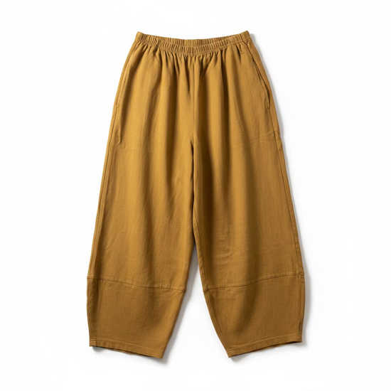 A pair of loose, wide-leg pants in a mustard yellow or ochre color, hanging from a black hanger against a textured off-white wall.