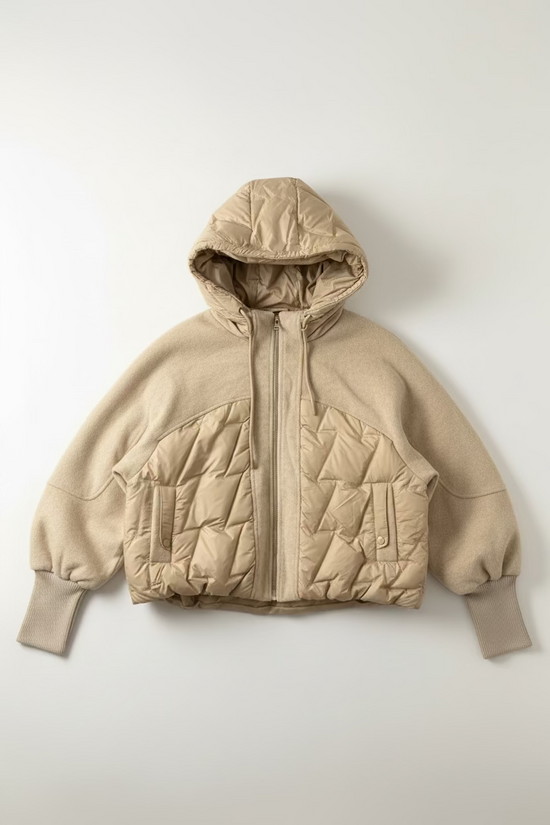 A beige hooded puffer jacket with quilted panels and a front zipper, hanging on a black wall hook against a textured off-white wall.