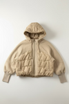 A beige hooded puffer jacket with quilted panels and a front zipper, hanging on a black wall hook against a textured off-white wall.