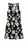 A sleeveless black dress with a white floral pattern, hanging on a black hanger against a textured off-white wall.